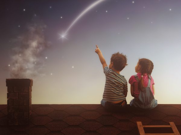 Two cute children sit on the roof and look at the stars. Boy and girl make a wish by seeing a shooting star.
